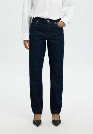 Persoon draagt donkerblauwe rechte jeans, wit overhemd met lange mouwen en knopen, en zwarte schoenen met puntige neus, staand met één hand in de zak.