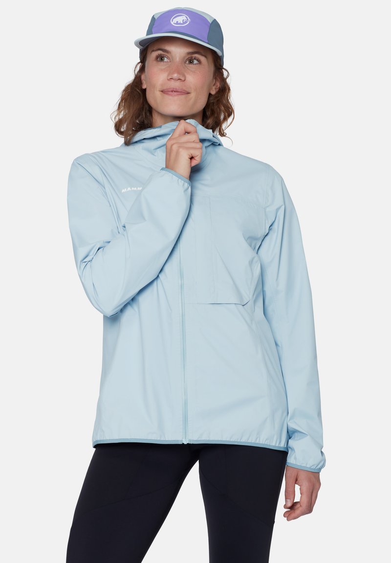 Frau in hellblauer Jacke mit Reißverschluss und lila-grauem Cap, schaut nach oben, hält den Jackenkragen, trägt schwarze Hose vor weißem Hintergrund.