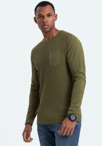 Maglietta a maniche lunghe verde oliva con texture a coste, dotata di tasca sul petto e scollatura tonda. Abbinata a jeans blu.