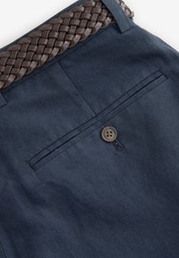 Pantalon bleu foncé avec une poche arrière boutonnée et une ceinture en cuir tressé marron passée dans les passants de ceinture.