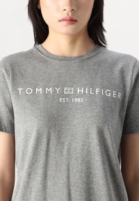 Šedé bavlněné tričko s kulatým výstřihem a krátkými rukávy. Na přední straně je nápis "TOMMY HILFIGER EST. 1985" bílým písmem.