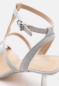 Ασημί γυαλιστερό πέδιλο με ψηλό τακούνι, λουράκι στον αστράγαλο και ασημί αγκράφα σε μπεζ πάτο, πάνω σε λευκό φόντο.