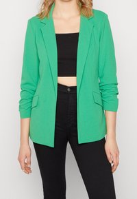 Blazer verde con un corte entallado, solapas con muesca, mangas largas y dos bolsillos delanteros, combinado con un top corto negro y pantalones negros.