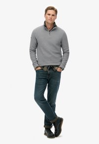 Grijze gebreide pullover met ritskraag, gedragen over een geruit shirt, in combinatie met blauwe jeans en zwarte veterschoenen, met opgerolde mouwen.