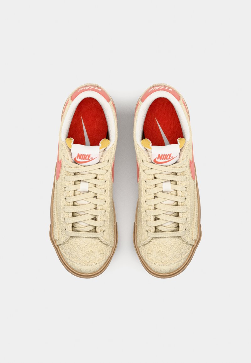 Baskets en suède beige avec une finition texturée, intérieur rouge et accents roses. Présentent des lacets classiques et une semelle en caoutchouc gomme. Marque Nike visible.