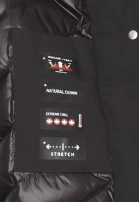 Giacca nera con interno lucido e isolante. Le etichette mostrano "Tessuto Hidra-neK", "Piuma Naturale", "Estrema Gelatezza" e icone per elasticità e ventilazione.