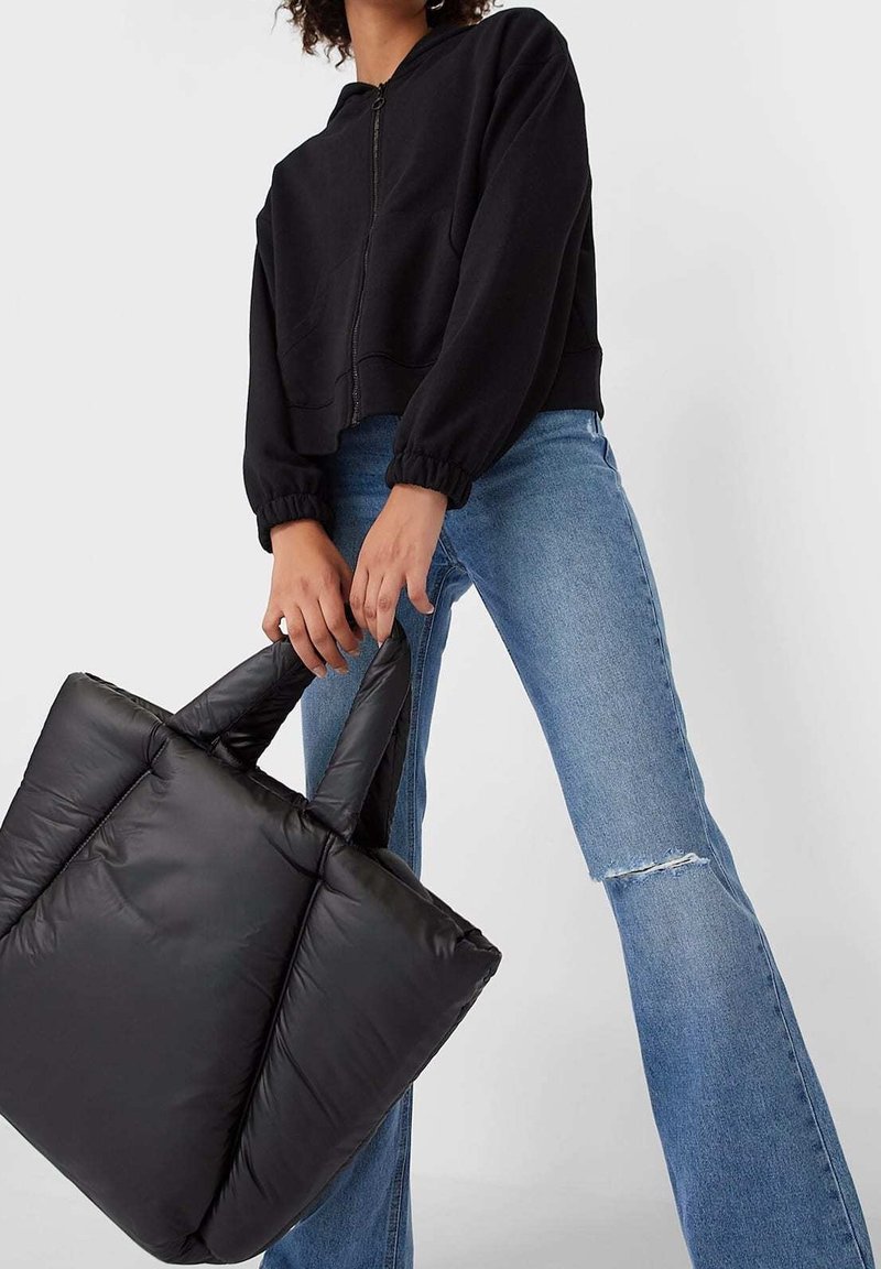 Personne portant une veste noire zippée et un jean bleu tient un grand sac fourre-tout noir matelassé par les deux poignées.