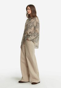 Genomskinlig beige blus med blommönster, knappstängning fram, vida manschetter; matchad med ljusbeiga vida byxor och bruna skor.