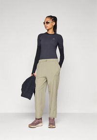 Sportovní outfit zahrnující černé tričko s dlouhým rukávem, světle šedé kalhoty a hnědé turistické boty. Součástí jsou sluneční brýle a tmavá bunda.