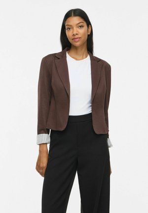Donna con lunghi capelli scuri indossa un blazer marrone sopra una maglia bianca e pantaloni neri a vita alta e gambe larghe su uno sfondo semplice.