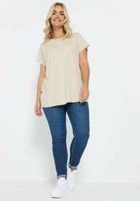 Beige kortærmet t-shirt med rund krave, parret med tætsiddende blå jeans og hvide sneakers. Outfittet har en afslappet, løstsiddende pasform.