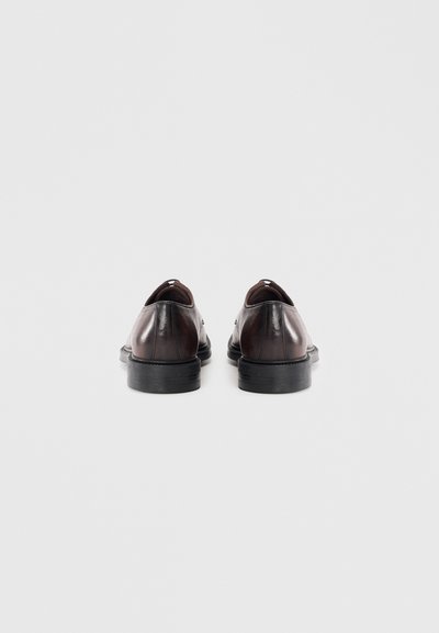 Zapatos de cuero marrón con un acabado suave, punta redondeada y suela baja de goma negra. La vista trasera muestra un ligero detalle de costura en el talón.