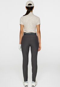 Donna in piedi di spalle, che indossa una polo leggera a motivi, pantaloni scuri aderenti, sneakers bianche, guanto da golf bianco e un cappellino beige con coda di cavallo.