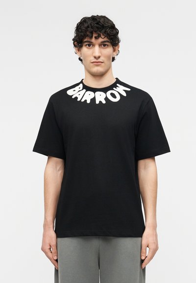 Άνδρας με σγουρά μαλλιά που φοράει μαύρο T-shirt με λευκή επιγραφή "BARRON" στη γιακά και γκρι παντελόνι, στέκεται μπροστά σε απλό φόντο.