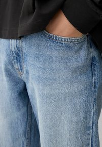 Oseba nosi svetlo modre jeans hlače, z eno roko v sprednjem žepu, črn dolg rokav majica pa je delno vidna.