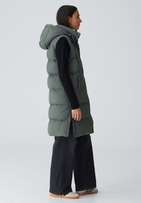 Puffige grüne Weste mit Kapuze, die ein langes Design und zwei seitliche Taschen aufweist, getragen über einem schwarzen Langarmoberteil und dunklen Jeans.