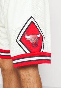 Pantalones cortos deportivos de malla blancos con acentos rojos, que presentan un parche en forma de diamante con una cabeza de toro y un dobladillo a rayas en blanco y negro.