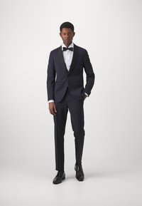 Marineblauer Smoking mit schwarzen Satinrevers, weißem Hemd und schwarzem Fliegenbinder; schmal geschnittene Hose und schwarze Lederschuhe. Minimalistisches Design.