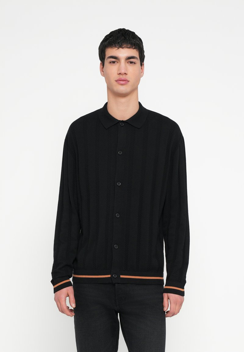 Fekete kötött kardigán függőleges bordázott mintával, nyolc gombbal, valamint kontrasztos narancssárga csíkkal az alsó szegélyen és az ujjtöveknél.