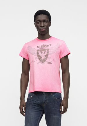 Jeune homme portant un t-shirt rose vif avec un blason d'aigle pixellisé et les mots vision et on life, associé à un jean bleu foncé.