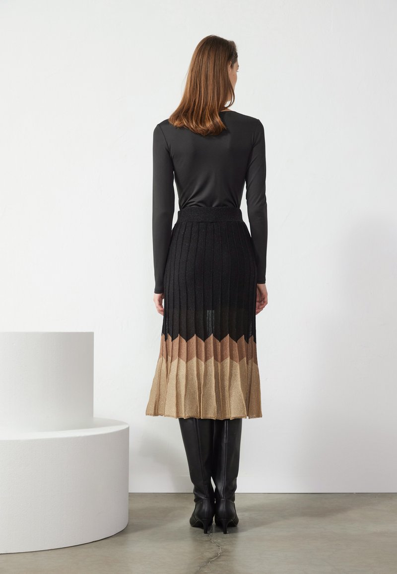 Schwarzes Langarmoberteil, kombiniert mit einem plissierten Rock, der einen Farbverlauf von Schwarz nach Hellbraun aufweist und aus strukturiertem Stoff gefertigt ist. Knielange schwarze Stiefel.