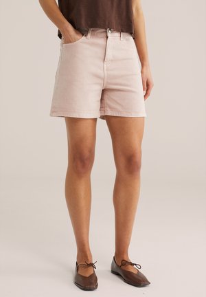 Kobieta ubrana w jasnoróżowe szorty dżinsowe, brązową bluzkę i brązowe pointy z wiązaniami wokół kostek, stojąca z jedną ręką w kieszeni.