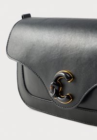 Schwarze Lederhandtasche mit abgerundetem Klappverschluss und schwarzem Harz-Verschluss in Form eines "C" mit Goldakzenten auf der Vorderseite, fotografiert vor weißem Hintergrund.