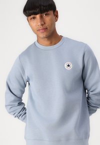 Lyseblå genser med rund hals, lange ermer og et sentralt logo-lapp med en stjerne, laget av et mykt, teksturert stoff.