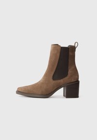 Bottine en suede marron avec un bout carré, des panneaux latéraux élastiques et un talon bloc. Le design comprend une boucle de traction pour un enfilage facile.