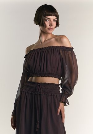 Kvinde med kort mørkt hår iført en mørk off-shoulder cropped top med gennemsigtige lange ærmer og matchende bukser med højt talje.