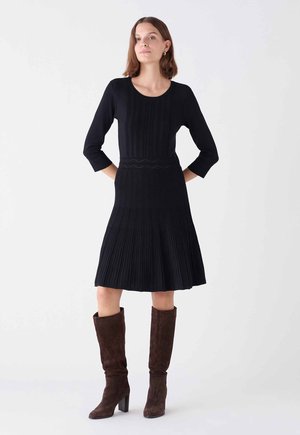 Robe en tricot noir avec une texture côtelée, manches 3/4, corsage ajusté et jupe plissée évasée. Accompagnée de bottes en daim marron montant jusqu'aux genoux.