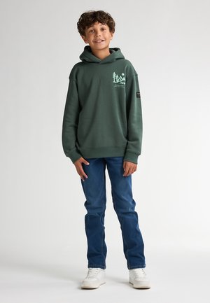 Sudadera verde oscura con un logo en la parte frontal, cuenta con un bolsillo tipo canguro y puños acanalados, combinada con jeans azules y zapatillas blancas.