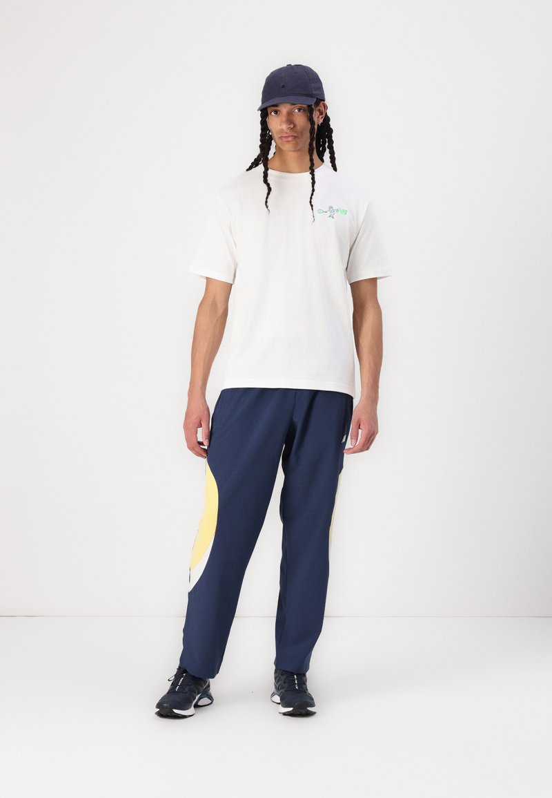 T-shirt en coton blanc avec un graphique vert, pantalon de jogging bleu marine avec des accents jaunes et blancs, et une casquette bleu marine. Chaussures de sport texturées.