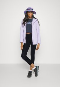 Lichtpaarse jas over een donkere crop top, zwarte leggings en grijze sneakers. Een kleurrijke, gemêleerde emmerhoed maakt de outfit compleet.
