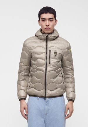 Jeune homme faisant face, portant une doudoune matelassée beige clair avec capuche et un jean bleu clair sur un fond uni.