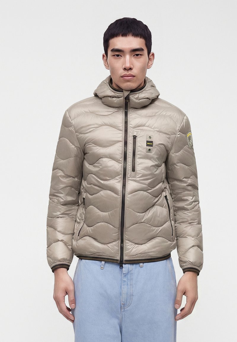 Jeune homme faisant face, portant une doudoune matelassée beige clair avec capuche et un jean bleu clair sur un fond uni.