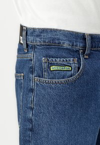 Jean en denim de couleur bleu moyen avec une coupe classique. Comprend un écusson à logo DC Shoes vert et jaune sur la poche arrière. Couture contrastante visible.