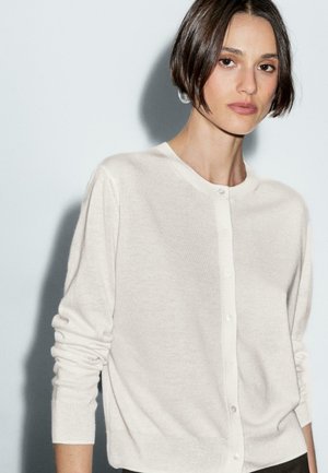 Eine Frau mit einem eleganten Bob-Haarschnitt trägt eine hellbeige Strickjacke und blickt direkt in die Kamera vor einem sanften grauen Hintergrund.