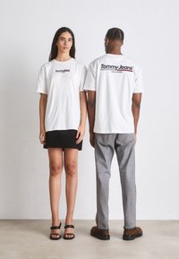 Camisetas de algodón oversized blancas con el logo "Tommy Jeans" en negro, llevadas por un modelo de frente y otro modelo mostrando la parte de atrás.