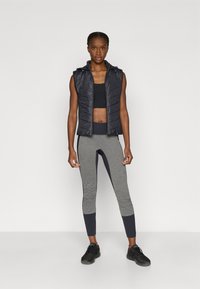 Veste sans manches noire, matelassée, avec fermeture éclair et capuche, associée à un crop top noir et des leggings gris avec des accents foncés et une coupe ajustée.