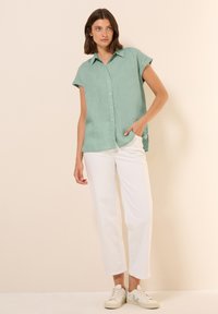 Camicia verde menta a maniche corte con bottoni, realizzata in tessuto leggero, abbinata a pantaloni bianchi a gamba dritta e sneakers chiare.