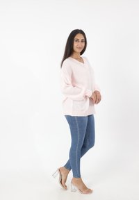 Hellrosa gestrickter Pullover mit V-Ausschnitt, überschnittenen Schultern und geripptem Saum. Kombiniert mit blauen Skinny Jeans und transparenten Blockabsatzsandalen.
