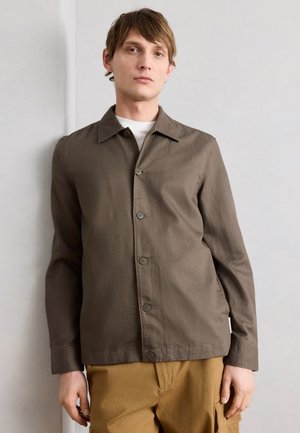 Ung mand iført en brun knapjakke over en hvid skjorte og sennepsfarvede bukser, stående mod en lysegrå væg.