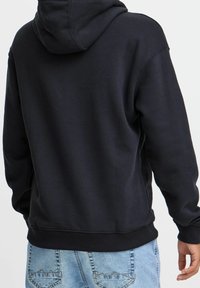Sudadera negra con capucha ajustable, puños y dobladillo acanalados, hecha de una tela suave. Lleva puesta sobre unos jeans azul claro con detalle en el bolsillo trasero.