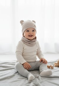 Cappellino per bambino color beige chiaro con orecchie da orsetto, scaldacollo beige, maglietta a maniche lunghe color crema, leggins grigi chiari, seduto su una coperta grigia con un giocattolo.