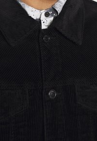 Chaqueta de pana negra con cuello de solapa, cierre de botones en la parte delantera y dos bolsillos en el pecho. Presenta un tejido de textura acanalada.