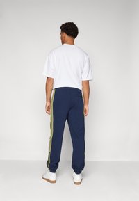 Calças de moletão azul-marinho com riscas laterais verde-lima, cintura elástica e bolso lateral. Usadas com uma t-shirt branca e ténis brancos.