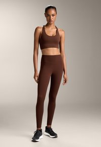Hnedá športová podprsenka a zodpovedajúce legíny vyrobené z hladkého, elastického materiálu, v kombinácii s čiernymi športovými topánkami. Jednoduchý dizajn a priliehavý strih.