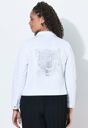 Frau mit lockigem Haar trägt eine weiße Jacke mit einem Strass-Tigerkopf-Design auf dem Rücken, kombiniert mit schwarzen plissierten Hosen.
