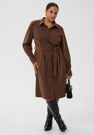 Femme portant une robe chemise marron ceinturée avec manches longues et des bottes noires montantes jusqu'aux genoux, tenant un sac à main noir sur un fond uni.